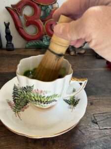 A hand whisks bright green matcha with a bamboo whisk in a vintage white teacup painted with green ferns, on a matching saucer, with a red and green Om carving and a small Ganesha statue on the wooden shelf behind.